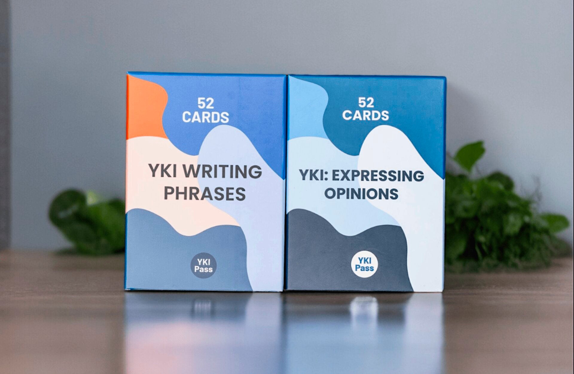 Two card packs stand side-by-side on a table. The left pack, labeled "YKI Writing Phrases," has an orange and blue design. The right pack, labeled "YKI: Expressing Opinions," has a blue and dark blue design. These tools are perfect for anyone looking to prepare for the YKI test and learn Finnish more effectively.